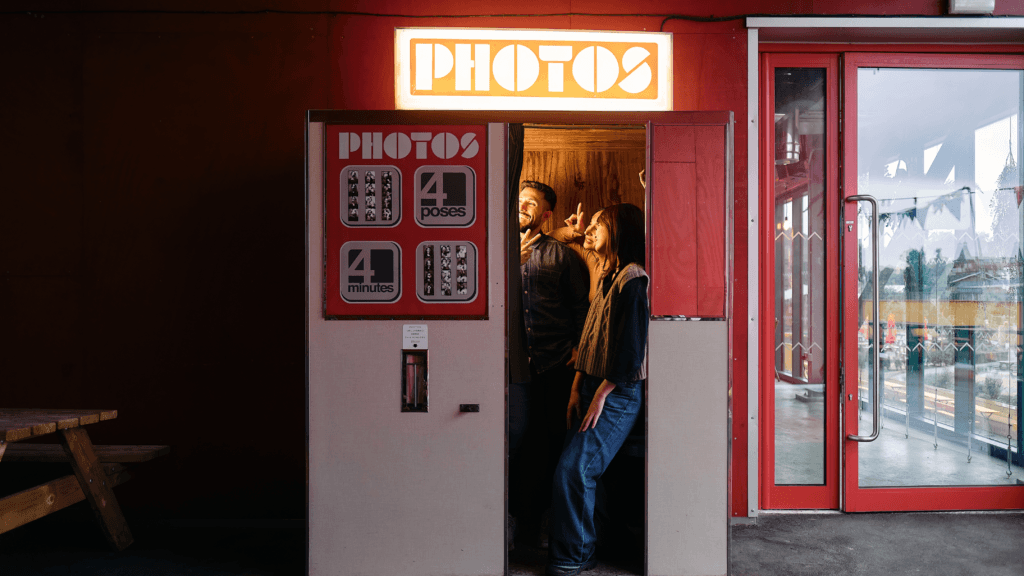 Ilustrasi Apa Bedanya Photobox dan Photobooth: Bilik photobooth klasik bertuliskan Photos dengan dua orang berpose di dalamnya, mencetak empat foto dalam empat menit